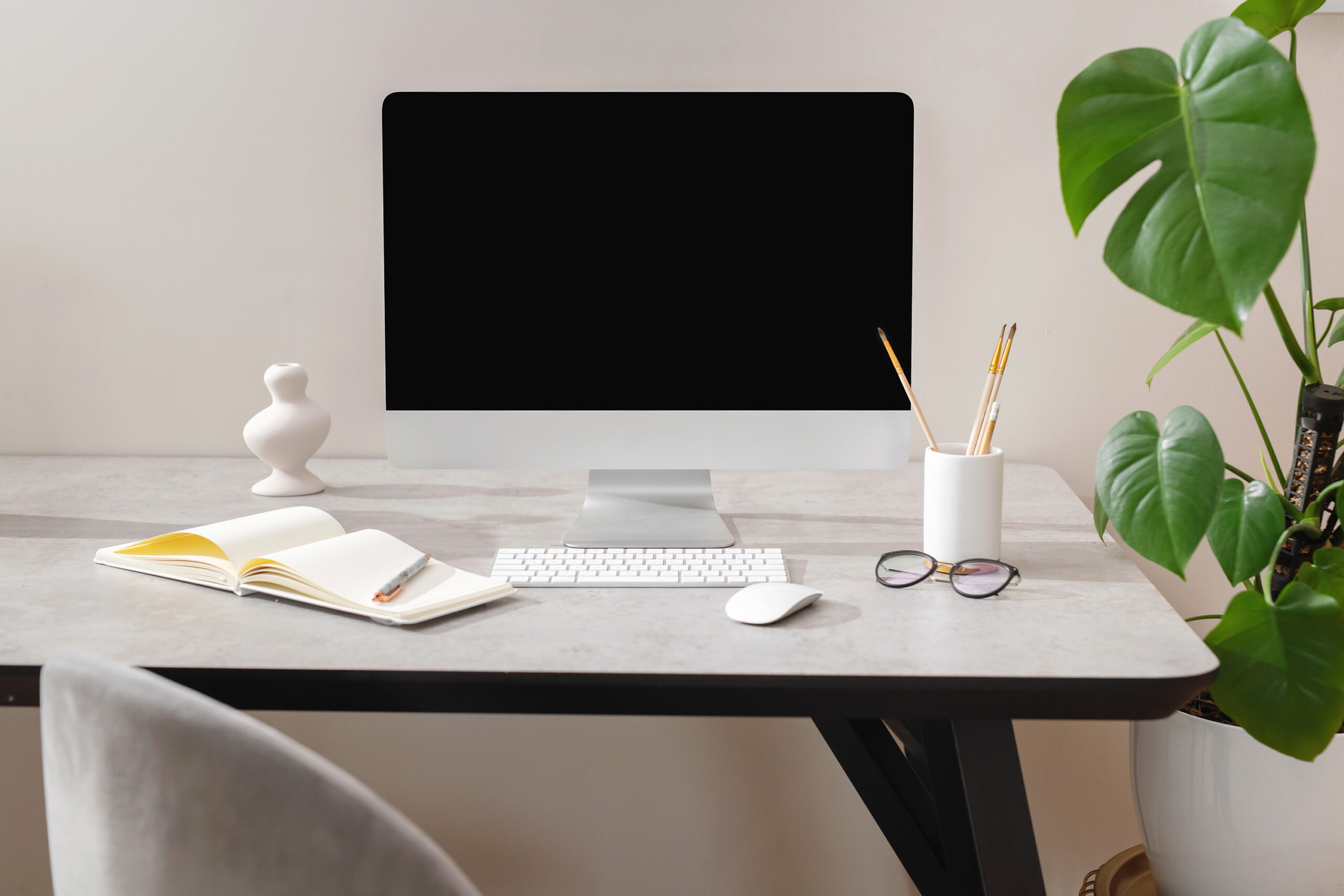 A stylish desk space with a desktop computer, glasses, notebook and pen, and pencil holder filled with pencils placed on top.